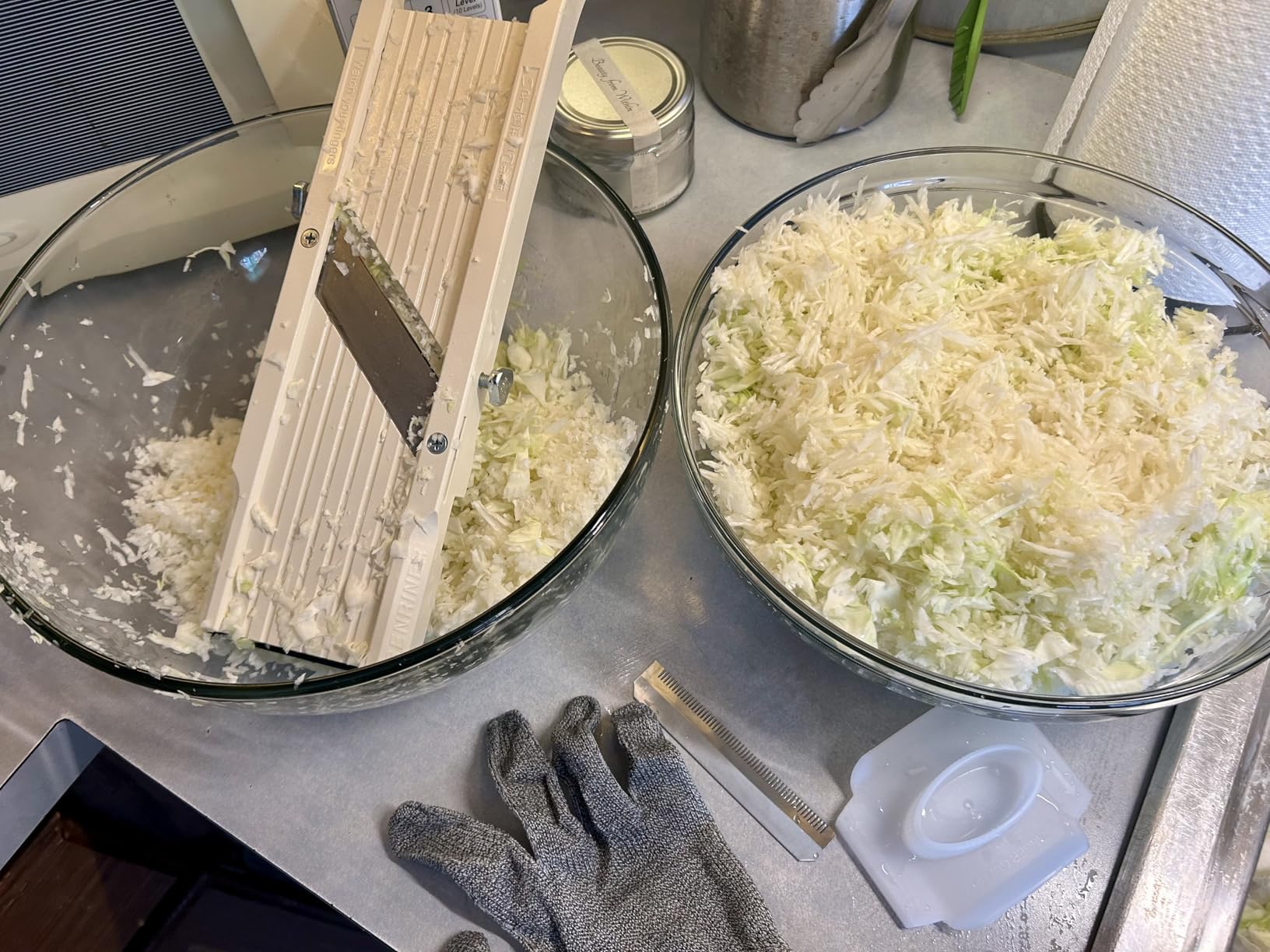 Two glass bowls of finely shredded cabbage beside a Benriner, with a cut-resistant glove and a comb blade on the counter